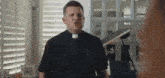 a priest is standing in front of a window in a living room talking to a woman .