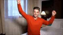 a man in a red shirt is sitting on a bed with his arms outstretched