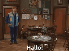 a man in a blue vest is standing in a room with tables and chairs and the words hallo on the table