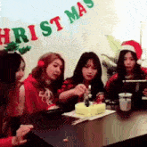 a group of girls are sitting around a table with a cake and a christmas banner behind them .