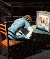 a man in a blue shirt is sitting at a table in front of a television