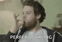 a man with a beard is blowing a kiss while sitting on a couch and says `` perfect darling '' .