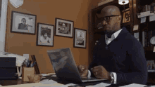 a man wearing glasses sits at a desk with a laptop in front of him