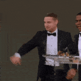a man in a tuxedo stands at a podium with another man