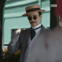 a man wearing a hat and sunglasses is standing in front of a building with hbo on the bottom right corner .