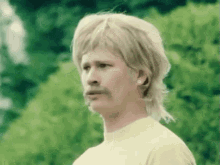 a man wearing a blonde wig and a mustache is standing in front of a green background .