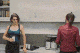 two women are standing in a kitchen talking to each other . one is wearing a red jacket .