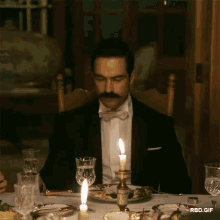 a man in a tuxedo sits at a table with candles lit