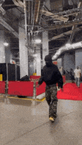 a man in a black hoodie and camo pants is walking down a red carpet in a building .