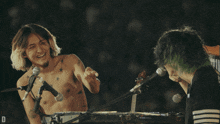 a man without a shirt is singing into a microphone while another man with green hair is playing a keyboard