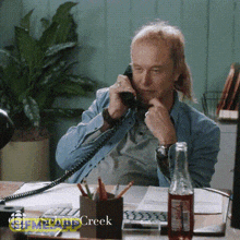 a man with a mullet is sitting at a desk talking on a telephone .