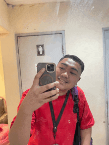 a man in a red shirt takes a selfie in front of a sign that says men 's restroom