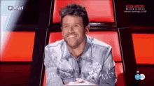 a man in a denim jacket is sitting in a red chair and smiling ..