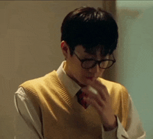 a young man wearing glasses , a yellow vest , and a tie is smoking a cigarette .