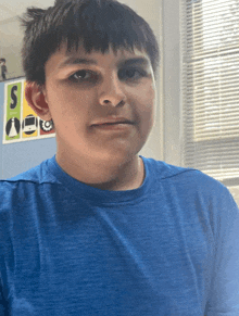 a boy wearing a blue shirt stands in front of a window with blinds