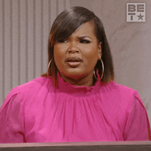 a woman wearing a pink shirt and hoop earrings stands in front of a be logo