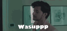 a man with a beard is standing in a room with the words wasuppp written on the screen .