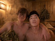 a man wearing glasses and a cross necklace stands next to another man in a sauna