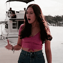 a woman in a pink crop top and jeans is standing in front of a boat in the water .