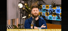a man with a beard stands in front of a microphone in front of a toni ryan wall