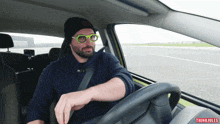 a man wearing glasses and a beanie is driving a car with thinkjules written on the bottom right