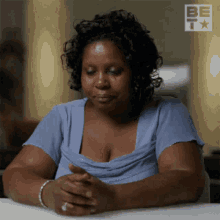 a woman in a blue shirt is sitting at a table with her hands folded in front of her face