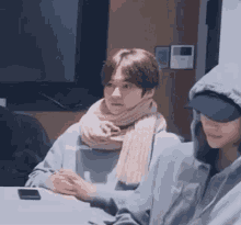 a man wearing a scarf and a hoodie is sitting at a table with another man .