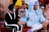 a group of drag queens are sitting on a couch and one of them is wearing a fur coat