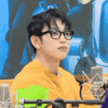 a young man wearing glasses and a yellow sweatshirt is sitting in front of a microphone .
