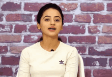 a woman wearing a white adidas shirt stands in front of a red brick wall