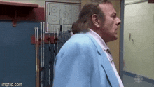 a man in a blue suit and tie is standing in a locker room with hockey sticks hanging on the wall .