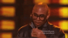 a bald man wearing glasses and a leather jacket covering his mouth with his hand .