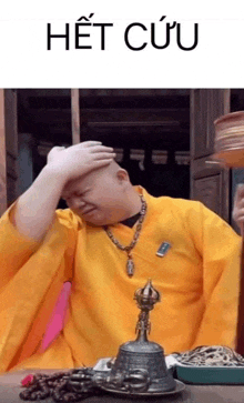 a man in a yellow robe is sitting at a table with a bell and a necklace .