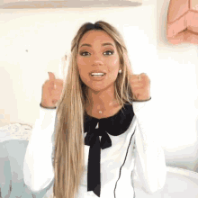 a woman is giving a thumbs up and wearing a white shirt with a black bow
