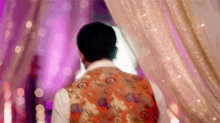 a man in a floral vest is standing in a room with a pink curtain .