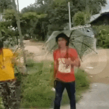a man in a red shirt is holding an umbrella while standing on a dirt road .
