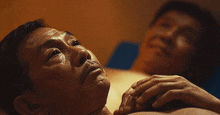a man with a beard is laying on a bed with another man in the background