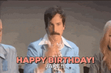 a man with a mustache is standing in front of a group of people and saying happy birthday .