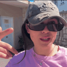a woman wearing sunglasses a hat and a necklace shows a peace sign