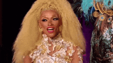 a drag queen with very long blonde hair is wearing a white and gold dress and earrings .
