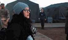 a woman wearing glasses and a beanie is standing in front of a group of soldiers
