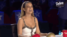a woman is laughing while sitting at a table in front of a screen that says deluxe