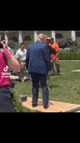 a man in a blue suit is standing on a wooden platform in the grass while a woman takes a picture of him .