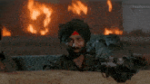 a man in a turban looks out from behind a wall with a fire in the background