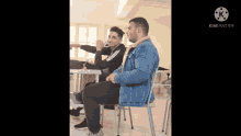 a man in a denim jacket sits on a chair next to another man in a black sweatshirt