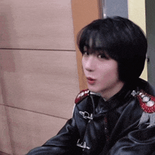 a woman wearing a black leather jacket with red shoulder pads is sitting in front of a wooden wall .