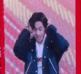 a man in a black jacket is standing in front of a red wall and holding his head .