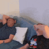 two men are sitting on a blue couch laughing and one has a mickey mouse shirt on