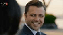 a man in a suit and tie smiles in front of a trt 1 logo