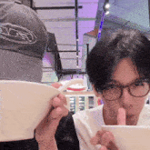 a man wearing glasses is eating from a bowl while another man wearing a hat with the letter e on it watches